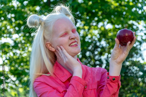 An albino girl in a red blouse against a background of green leaves and sunlight. she holds a red Apple in her hands and looks at it with distaste.