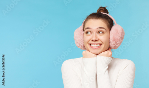 Young smiling girl in pink earmuffs, looking away, dreaming of something, isolated on blue background with copy space