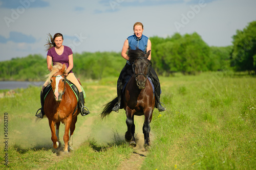 Two Caucasian horsewomen are riding along the rural road. The horses hooves are throwing up the dust-clouds.