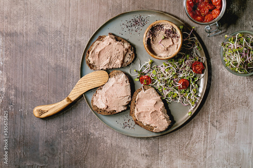 Sandwiches and ceramic bowl of homemade chicken liver pate with wooden knife, sun-dried tomatoes and green sprout salad on gray plate over dark texture background. Flat lay, space