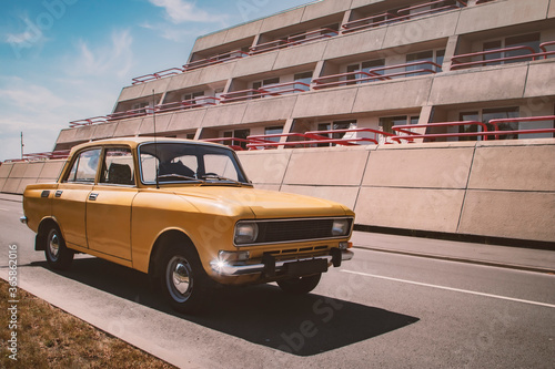 A nostagic vintage photo of a yellow retro classic soviet russian AZLK Moskvich 2140 car wearing an old black blank licence plate parked in front of a 1980s concrete building on a sunny summer day