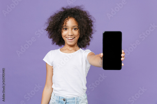 Smiling little african american kid girl 12-13 years old in white t-shirt isolated on violet wall background studio portrait. Childhood lifestyle concept. Hold mobile phone with blank empty screen.