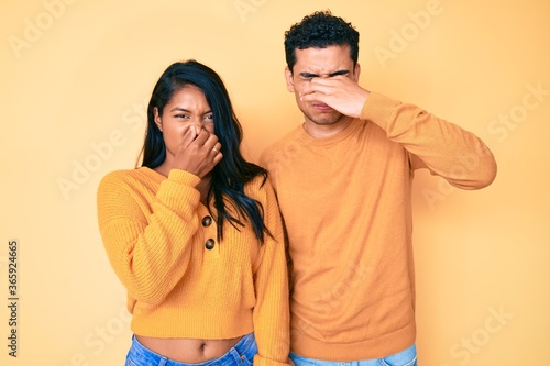 Beautiful latin young couple wearing casual clothes together smelling something stinky and disgusting, intolerable smell, holding breath with fingers on nose. bad smell