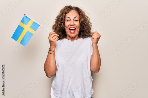 Middle age beautiful tourist woman holding swedish flag over isolated white background screaming proud, celebrating victory and success very excited with raised arm