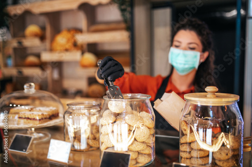 Beautiful young female worker with protective mask on face working in bakery.