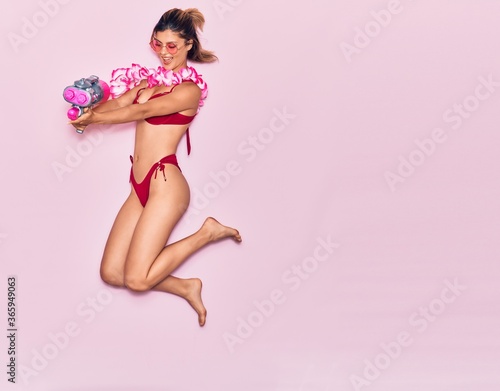Young beautiful girl on vacation wearing bikini and hawaiian lei smiling happy. Jumping with smile on face holding water gun over isolated pink background