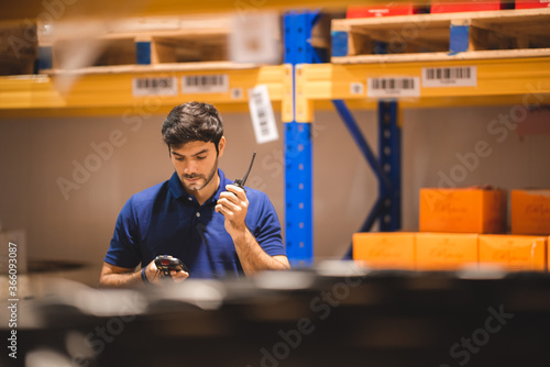 Male dispatcher, warehouse worker checking packages while using walkie-talkie in distribution warehouse storage.