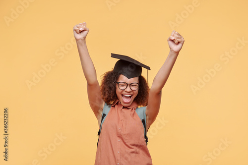Hilarious emotional Afro-American student girl in academic cap keeping eyes closed while celebrating her success