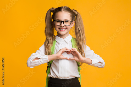 Smiling schoolgirl gesturing heart shape with fingers on yellow background
