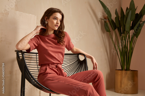 Beautiful brunette woman natural makeup wear fashion clothes casual dress code office style total pink blouse and pants suit, romantic date business meeting armchair interior stairs flowerpot.