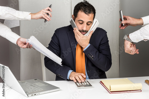 Manager i giacca e cravatta seduto nella postazione di lavoro del suo ufficio è circondato da mani e impegni e fa tante cose contemporaneamente