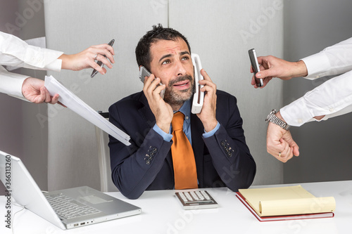 Manager i giacca e cravatta seduto nella postazione di lavoro del suo ufficio è circondato da mani e impegni e reagisce in maniera stressata mentre risponde al telefono