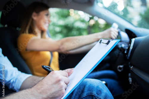 Close up view of driving instructor holding checklist while in background female student steering and driving car. Acquiring driver's license.