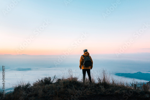 Back person contemplating an immense landscape covered with fog from the top of a mountain at sunrise. Traveller escaping in solitude into the wilderness. Connection to nature to the great outdoors.