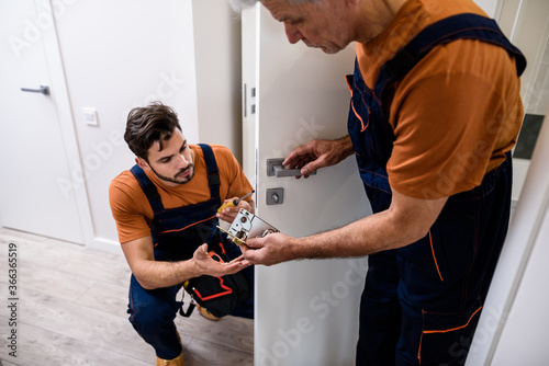 Two locksmith, repairmen, workers in uniform installing, working with house door lock using screwdriver. Repair, door lock service concept