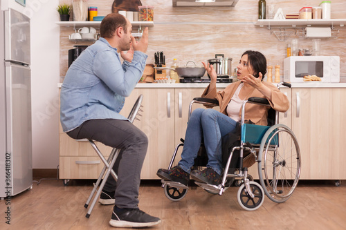 Disabled wife in wheelchair shouting at husband in kitchen. Disabled paralyzed handicapped woman with walking disability integrating after an accident.