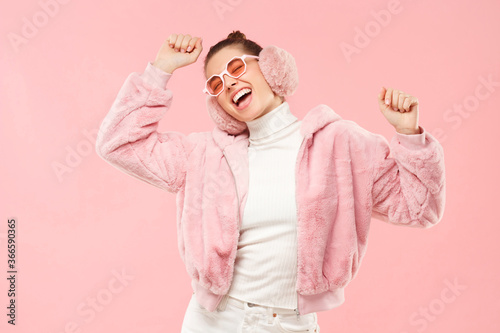 Young excited girl wearing warm furry coat, earmuffs and colored eyeglasses, moving arms to sounds of music, isolated on pink background