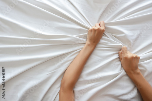 Top view young sexy woman hands pulling white sheets in ecstasy in hotel. Cute girl doing sign orgasm on white bed (sex and erotic concept for advertisement)