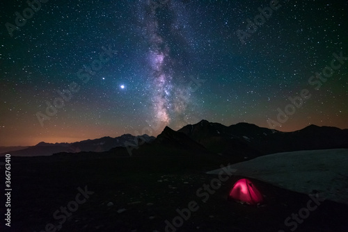 Milky way galaxy stars in the night sky over the Alps, illuminated camping tent in foreground, snowcapped mountain range, astro photography stargazing