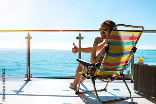 Attractive beautiful smiling woman with long hair and sunglasses sitting in a chase lounge on the balcony with seascape view and looking at the camera raised finger.