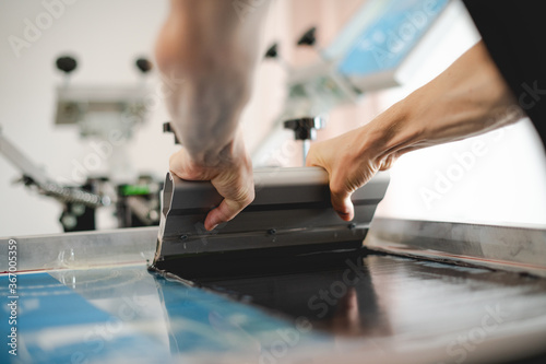 Selective focus of craftsman with squeegee working on screen printing machine in workshop 