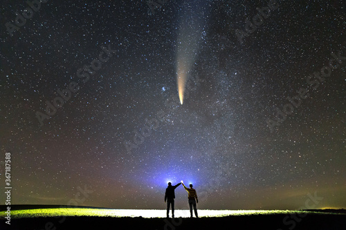 Small silhouettes of two scientists with flashlight on heads pointing bright beam of light on starry sky with C/2020 F3 (NEOWISE) comet with light tail. Space exploration concept.