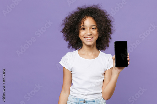 Smiling little african american kid girl 12-13 years old in white t-shirt isolated on violet wall background studio portrait. Childhood lifestyle concept. Hold mobile phone with blank empty screen.