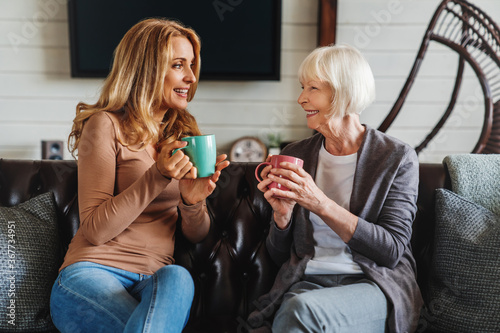 Smiling mature mother and adult daughter on sofa in living room talking and drinking tea or coffee