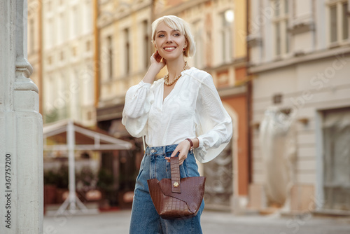 Street style photo of happy smiling fashionable woman wearing trendy white blouse, high waist jeans, holding brown faux croco leather textured bag. Model posing in street of European city. Copy space 