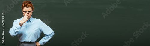 horizontal concept of strict teacher touching eyeglasses while standing with hand on hip near chalkboard