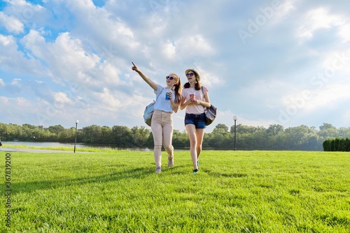 Two teenage girls walking together on the lawn with drinks on sunny summer day