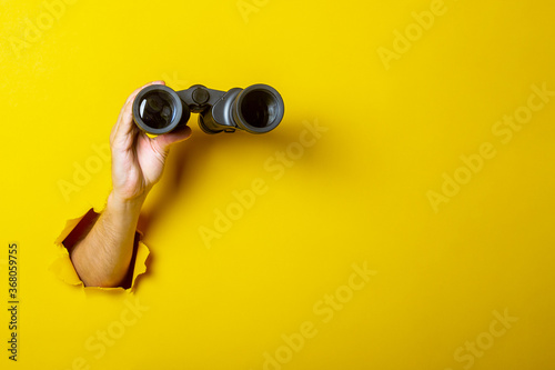 Female hand holds black binoculars on a yellow background. Looking through binoculars, journey, find and search concept