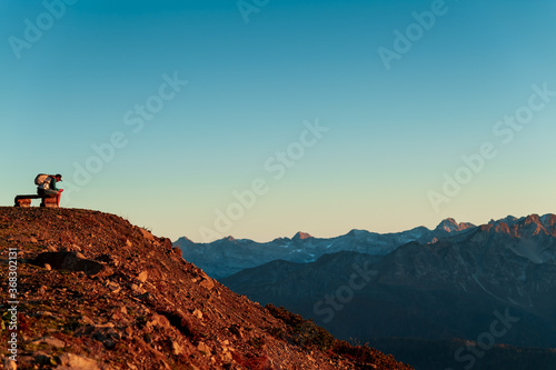 A guy in a cap and with a large backpack is resting on a bench overlooking a beautiful sunset in the mountains.