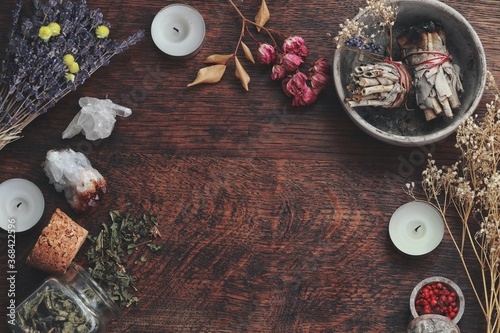 Dark brown wooden table background for text, decorated with witchy wiccan items to look esoteric and occult. Free writing space in the middle. Dried herbs, crystals, sage smudge sticks candles flowers