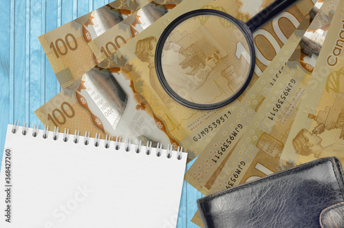 100 Canadian dollars bills and magnifying glass with black purse and notepad. Concept of counterfeit money. Search for differences in details on money bills to detect fake