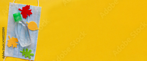 On the protective mask lies a sanitizer surrounded by felt autumn colored leaves on a yellow background. Flat lay. Top view.