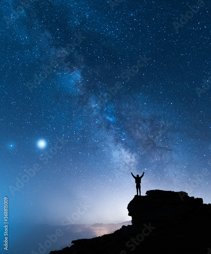 Man and the Universe. A person is standing on the top of the hill next to the Milky Way galaxy with his hands raised to the air.
