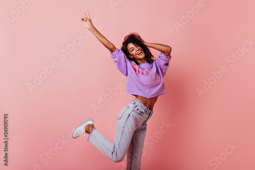 Shapely jocund girl dancing on rosy background. Good-humoured african lady jumping with smile.