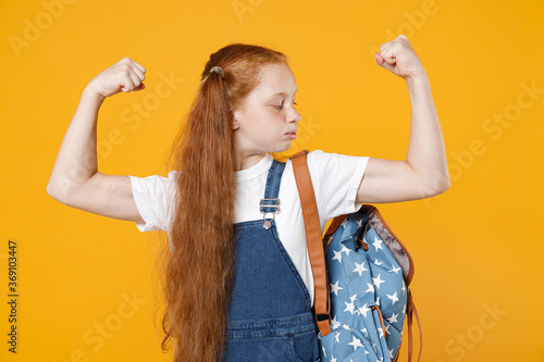 Strong young school teen girl 12-13 years old pony tails white t-shirt blue denim uniform backpack showing biceps muscles isolated on yellow background children studio portrait Kids education concept