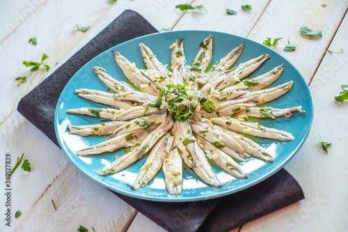  Pickled anchovies with garlic and parsley marinated in vinegar in a round wooden plate on a white wooden table. Typical Spanish snack. 