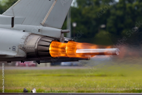 Afterburners glowing on an air force fighter jet aircraft as it speeds down the runway of an air base.