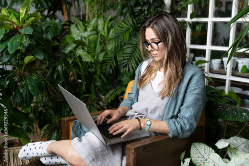 Concentrated female gardener in eyeglasses wear linen dress, sitting on chair in green house, working on laptop surrounded by exotic plants. Freelance