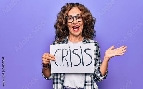 Middle age beautiful woman holding paper with crisis message over purple background celebrating achievement with happy smile and winner expression with raised hand