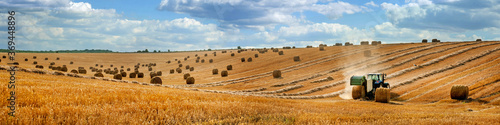 large panorama of a field with bales of straw, a tractor with a baler harvesting straw