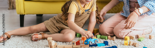 cropped view of babysitter near child playing with toy car near multicolored blocks on floor, horizontal image