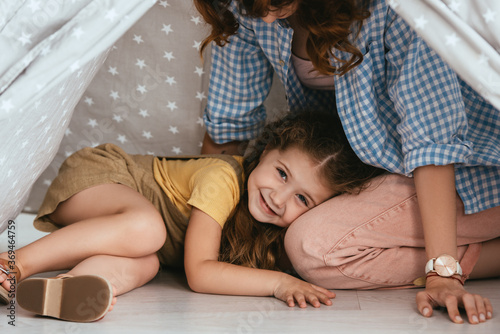 cropped view of babysitter and happy child looking at camera from play tent