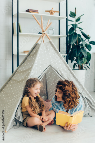 smiling babysitter looking at laughing child while holding book in kids wigwam
