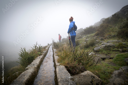 Two people hiking in the mountains in a foggy and rainy day. Girl with blue jacket and umbrella and man with red jacket.