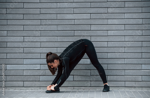 Young sportive girl in black sportswear outdoors doing stretching near gray wall