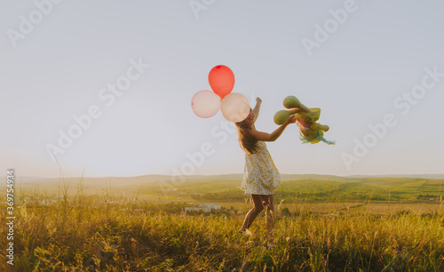 Side view portrait of a young girl playing with balloons and a doll on a hill backlit by sunset light in a rural setting